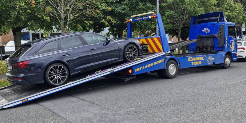 Blue Camel Tow flatbed recovery truck loading a dark grey Audi estate on a Dublin street, CAMEL TOW and 087 465 0060 on the truck